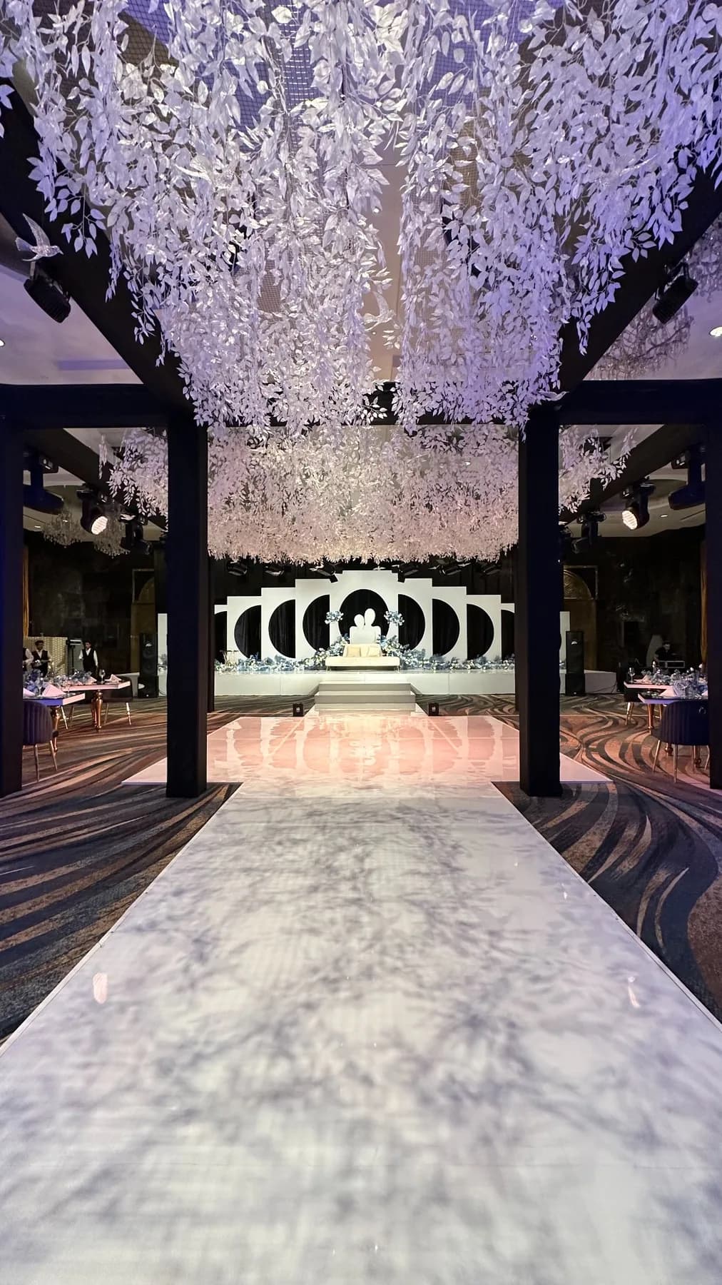 A pale aisle at Raffles Dubai beneath a dense canopy of white hanging foliage, framed by black rectangular supports