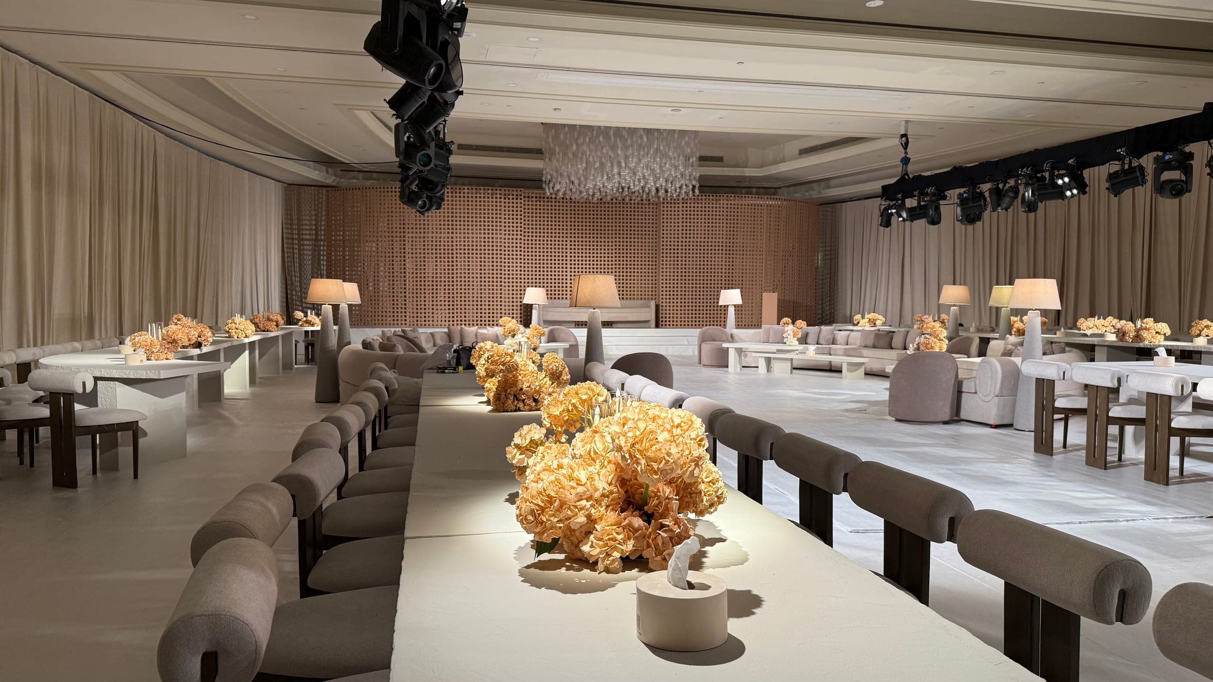 A long table with apricot hydrangeas leading towards a lattice wall, lounge seating and a suspended crystal installation, with lighting on a truss overhead