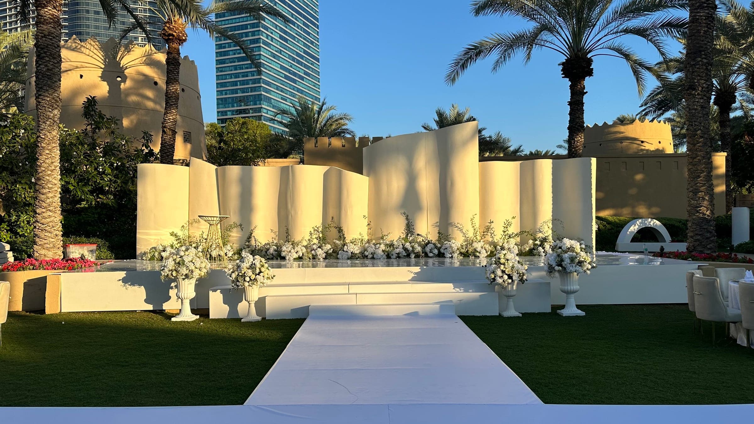 An outdoor stage of curved ivory panels lined with white florals, reached by a white aisle across the lawn among palms