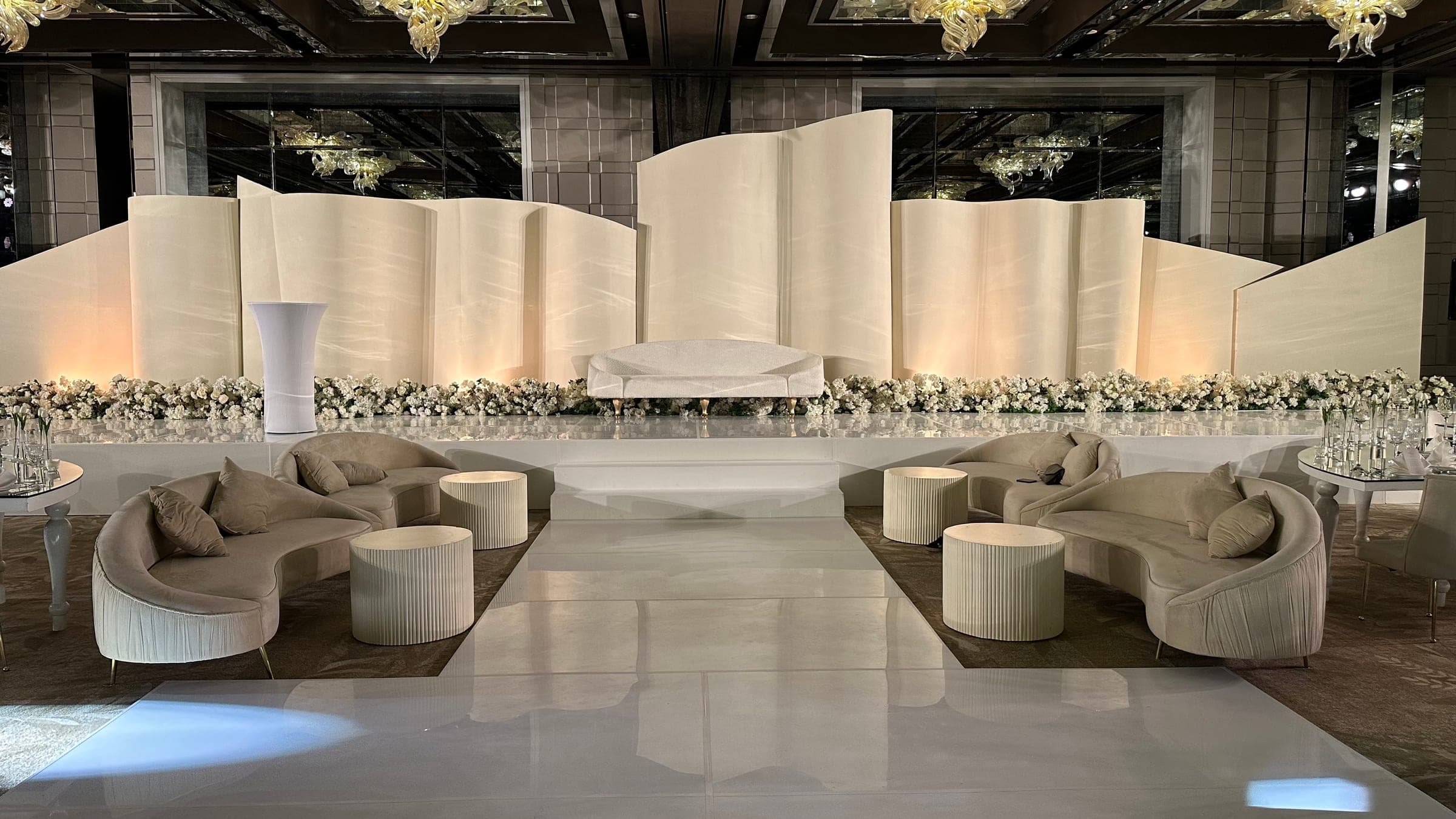 A stage of folded ivory panels with a line of white florals and a central sofa, lounge seating on either side of a glossy aisle