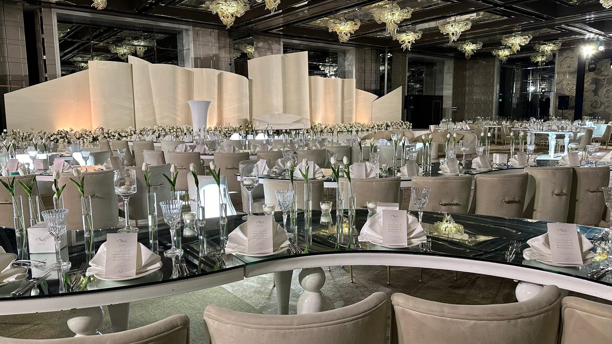 Mirrored tables with glassware and tulips before the ivory panel stage, beneath floral installations hanging from the ceiling