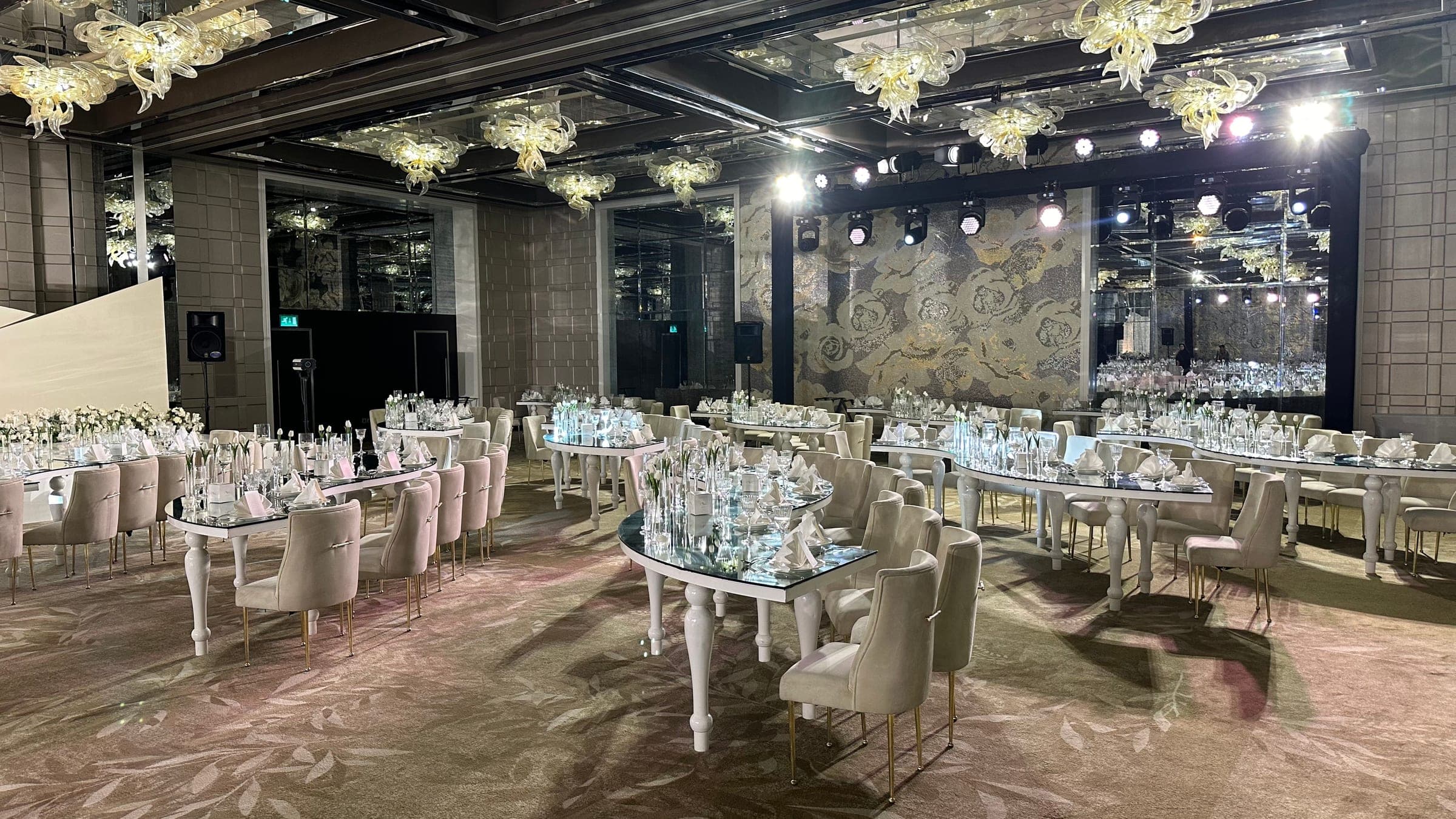 The room from the side: curved mirrored tables, floral installations across the ceiling and lighting on a truss