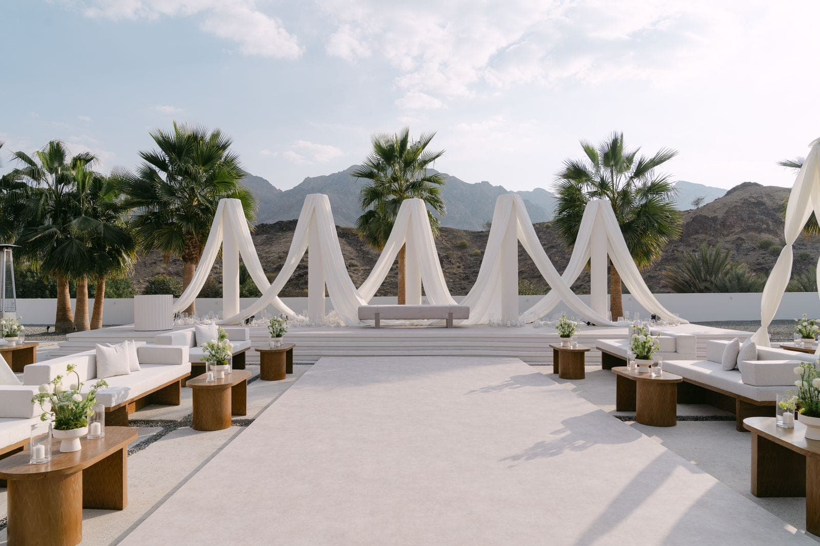 Lounge seating and wooden side tables along a pale aisle leading to arches of draped white fabric, with palms and mountains behind