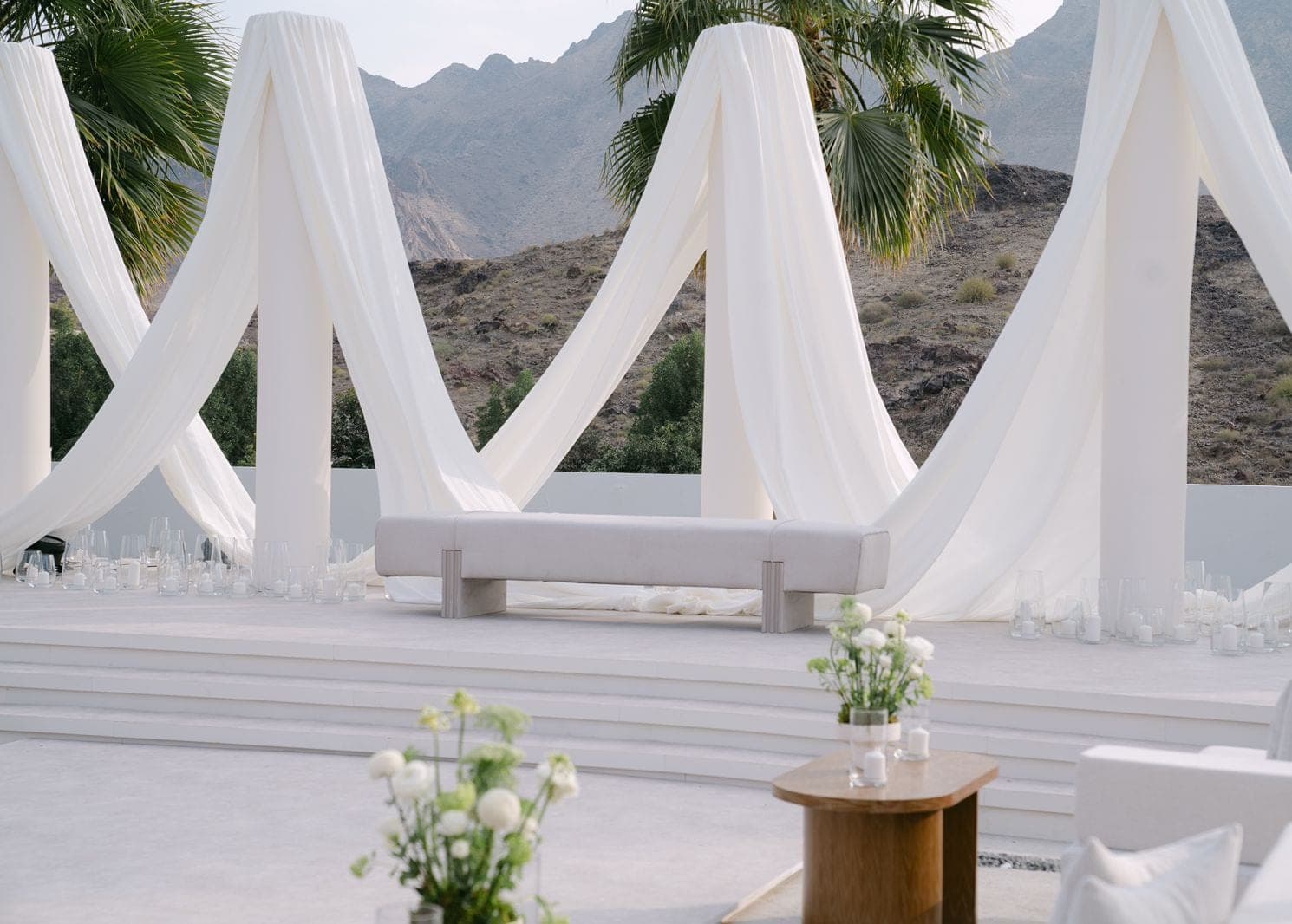 A low bench on a white platform framed by draped white fabric, with candles in glass and white flowers