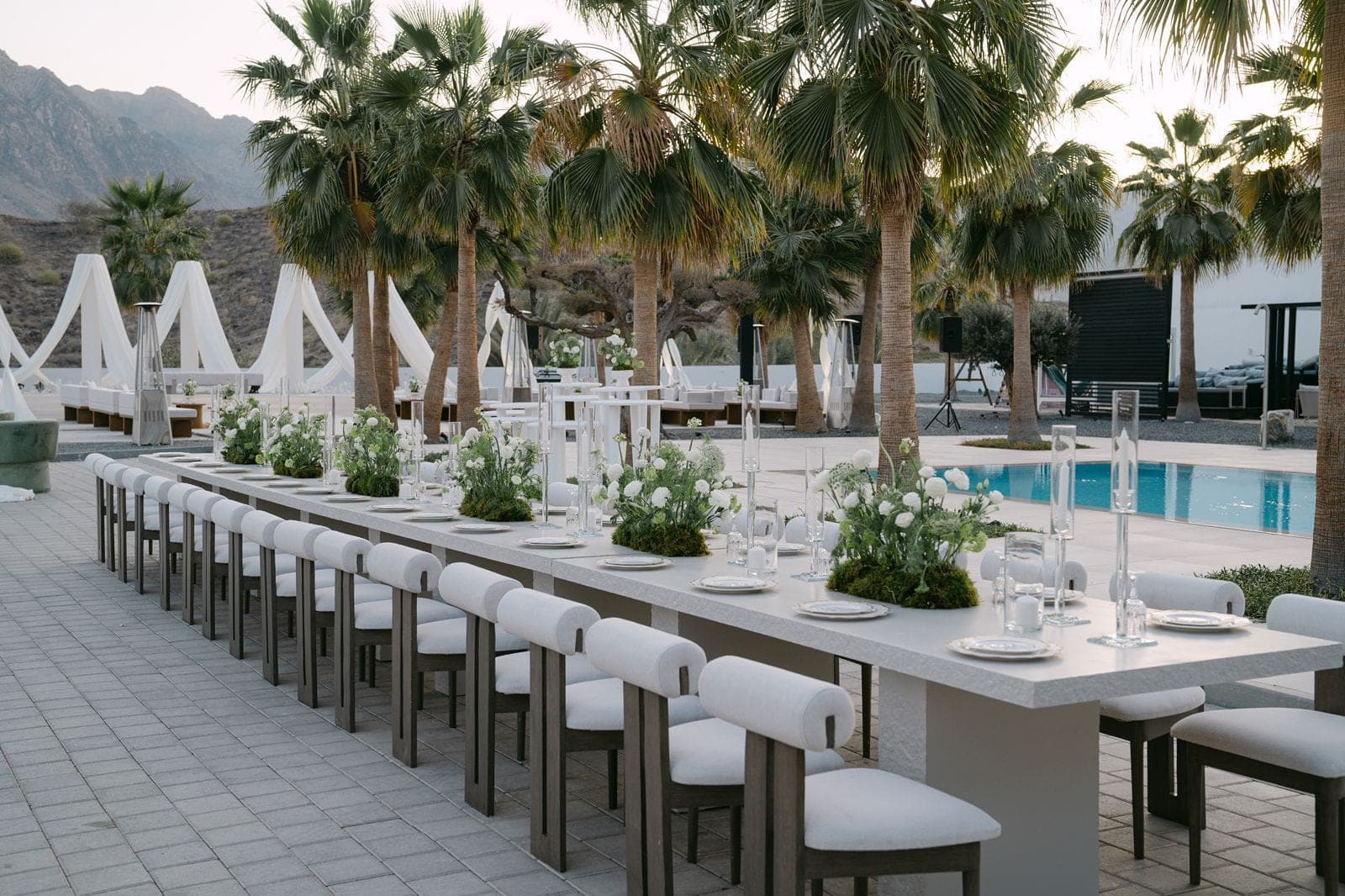 A long table beside a pool, set with white florals and tall glass candle holders, under palms
