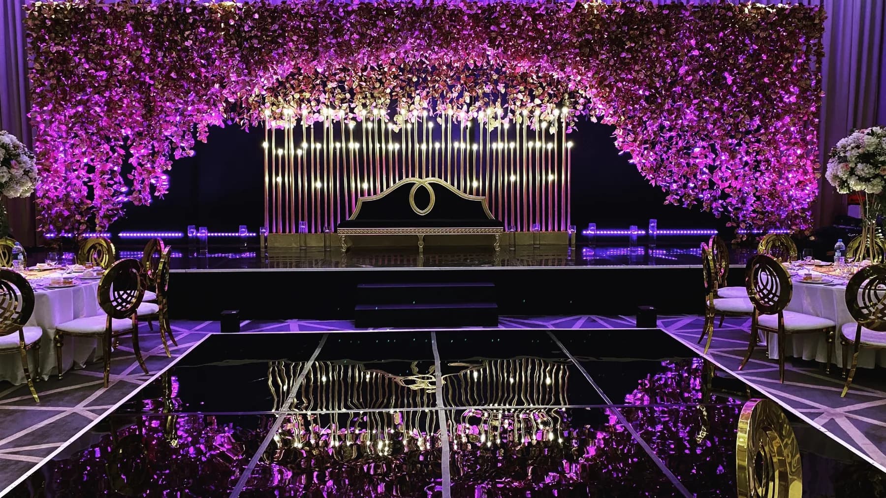 A Sheraton Grand Dubai ballroom with a glossy black dance floor leading to a stage framed by dense purple florals and vertical warm lights