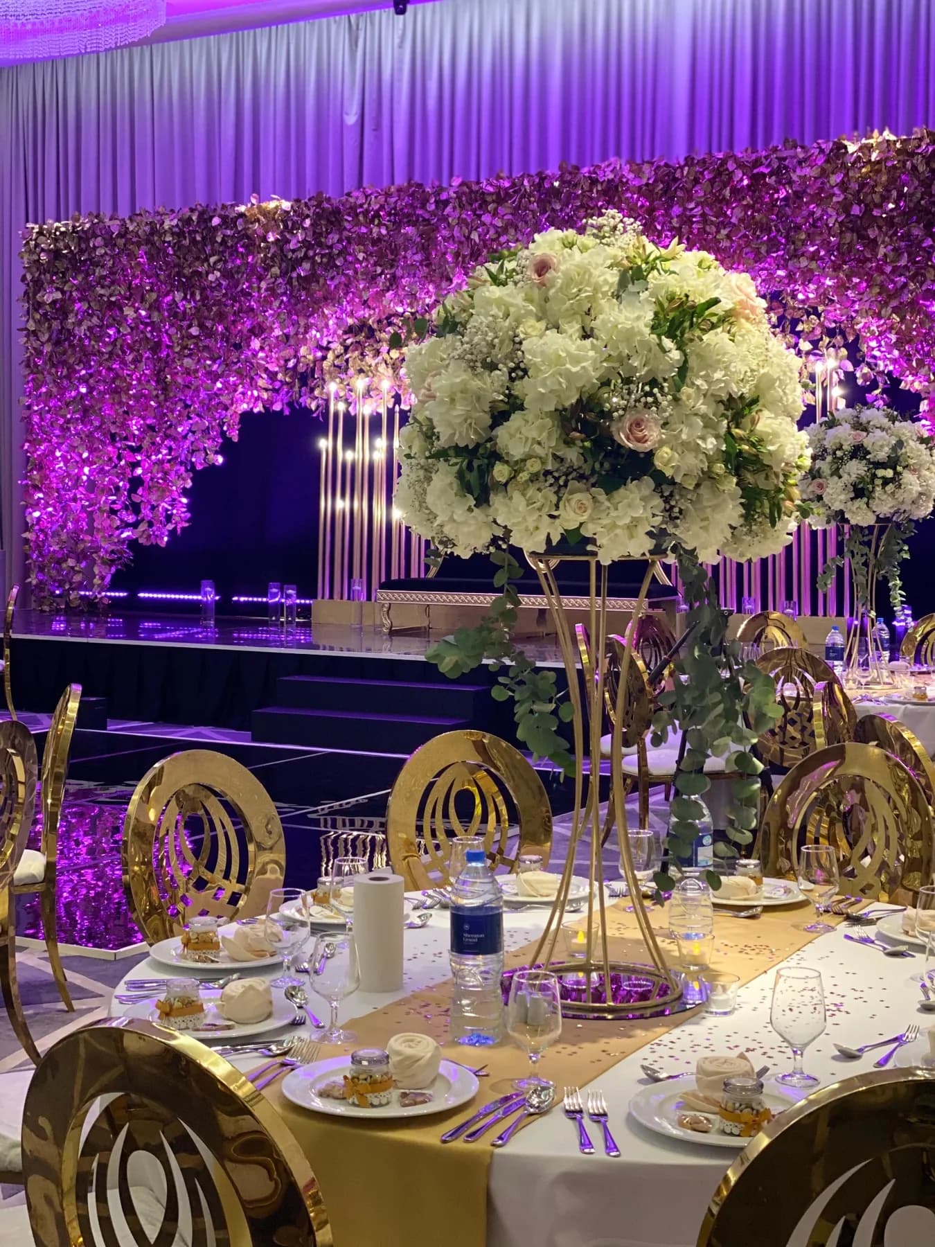 A round ballroom table with a tall white floral centerpiece and gold chairs, with the purple floral stage behind