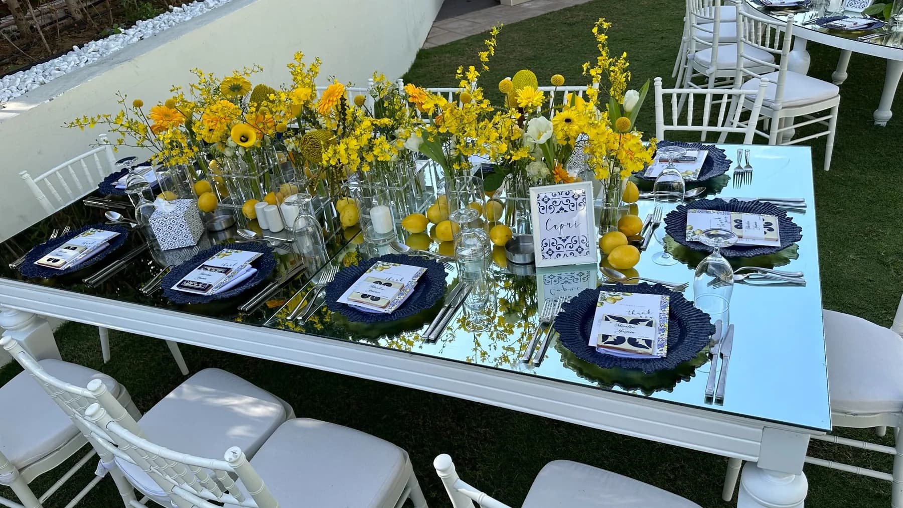 A mirrored outdoor dining table at Address Montgomerie styled with yellow florals, lemons, blue-and-white tableware and white chairs