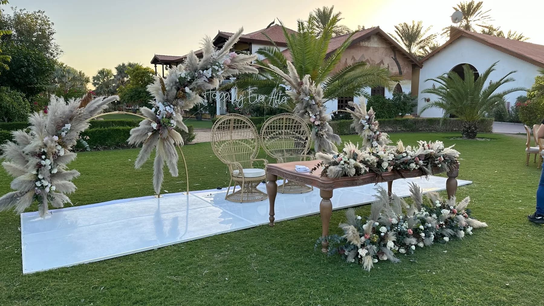 An outdoor wedding setting at Meliá Desert Palm with a white aisle, woven peacock chairs, pampas florals and a wooden signing table on the lawn