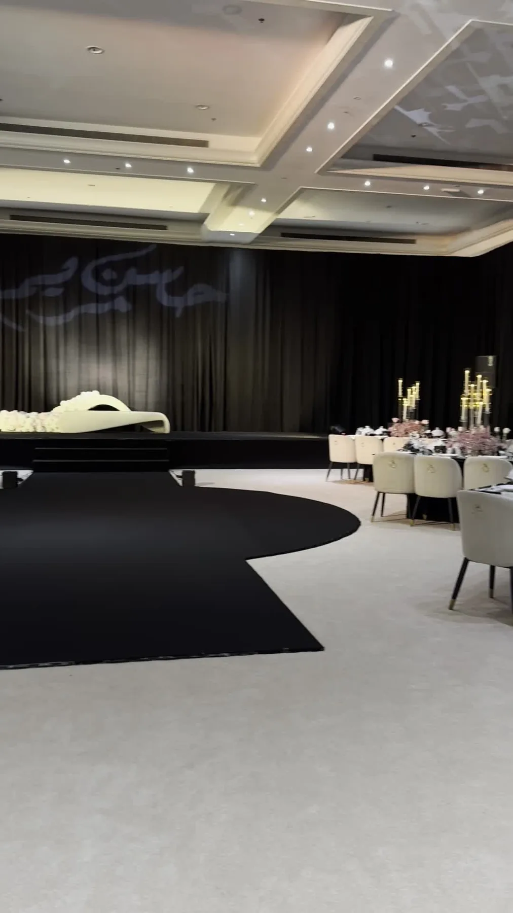 Al Dana Hall with a sweeping black floor treatment, black wall draping, round dining tables and a light-toned stage at the far end