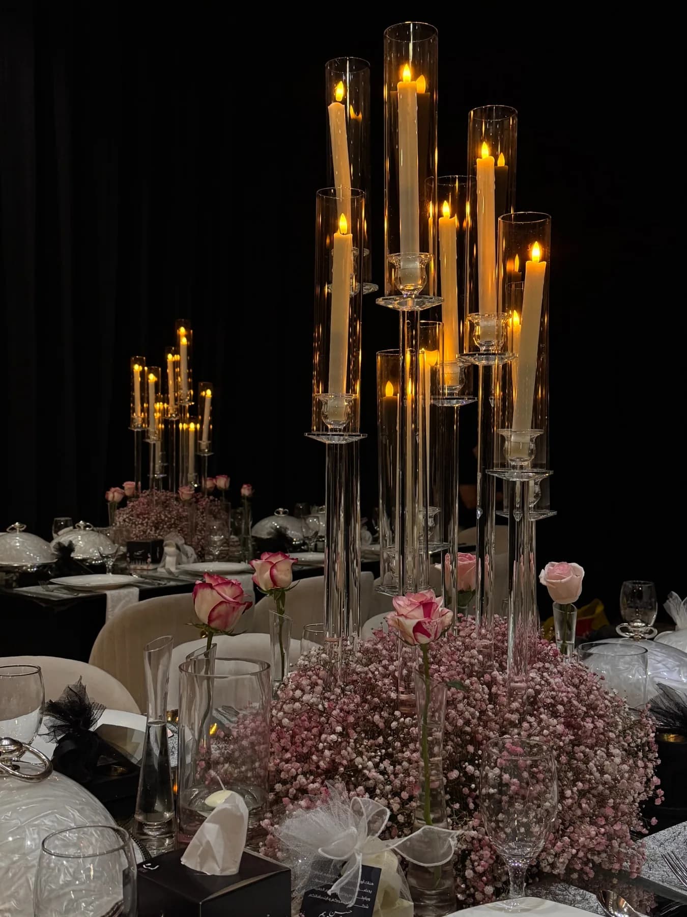 A tall glass candle centerpiece at Al Dana Hall rising above pink baby's-breath florals on a dining table